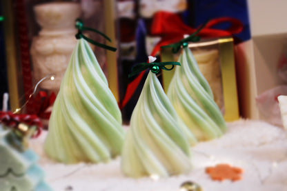 Drei handgefertigte grüne Weihnachtsbaum-Kerzen mit Schleifen und Glöckchen auf festlich dekoriertem Tisch.